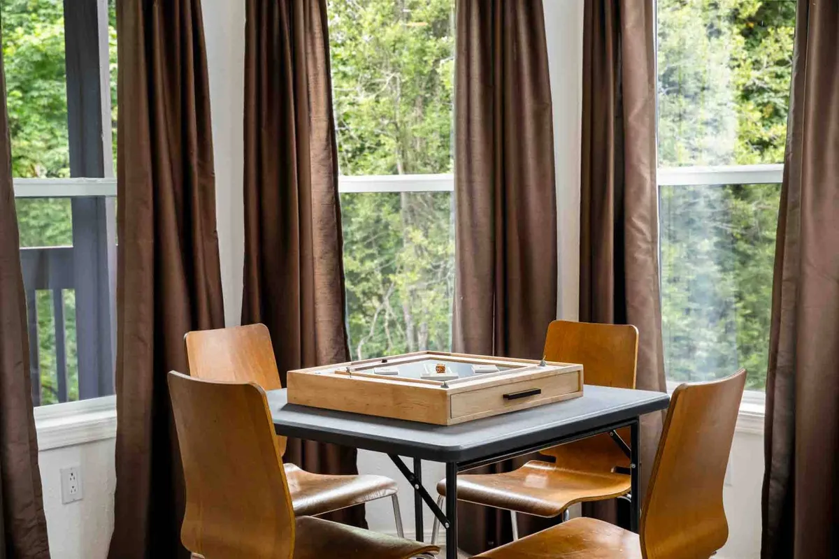 A cozy dining or game area with a wooden game board set up on the table, surrounded by chairs and soft curtains. The space is warm and inviting, suggesting an ideal spot for family board games and casual gatherings.