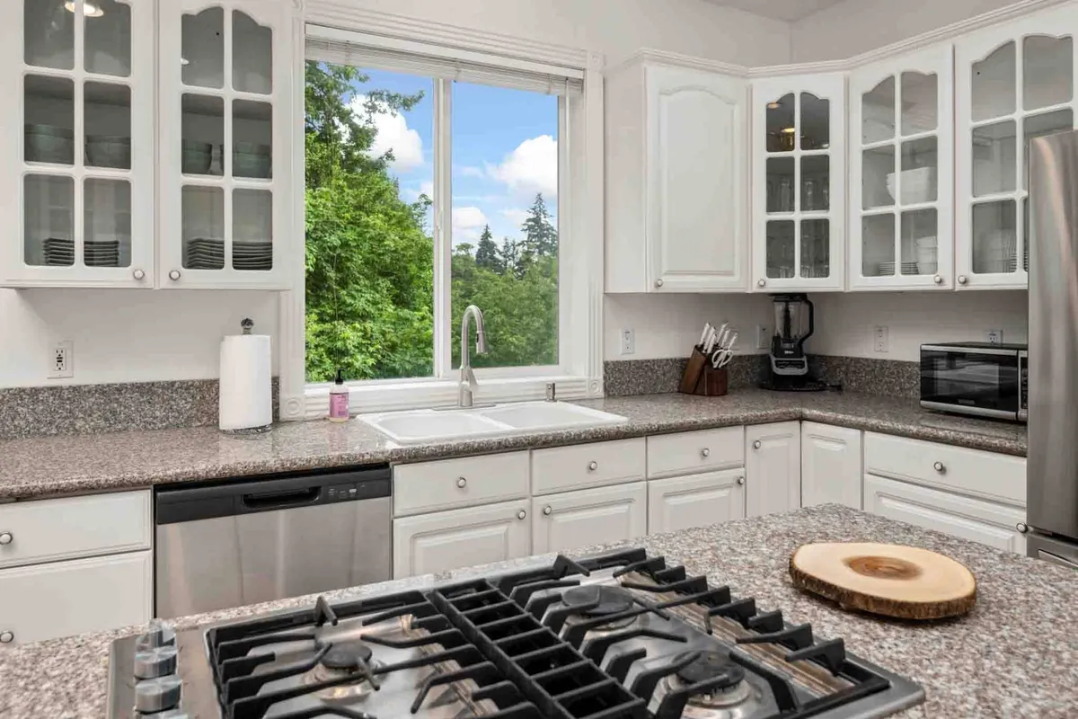 A kitchen with granite countertops, stainless steel appliances, open shelving, and a large window that frames an outdoor view. The room is sunlit, welcoming, and ready for morning coffee or group gatherings before heading out.