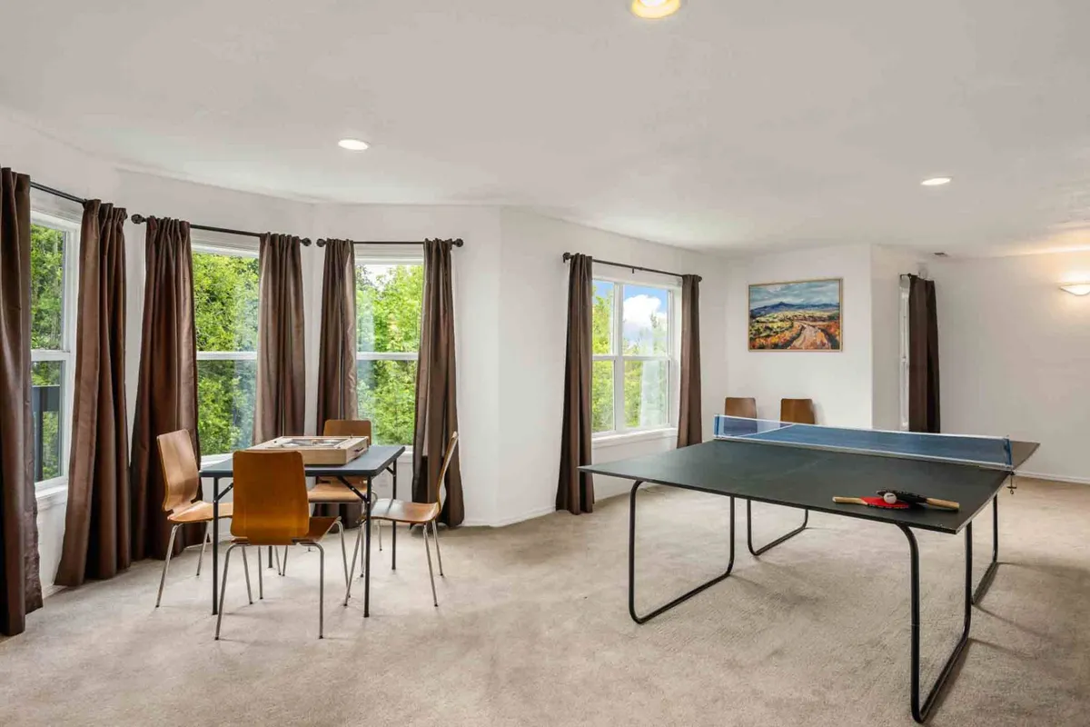 This stylish interior highlights a modern game area with a prominent ping pong table, a dining table for board games or snacks, expansive windows bringing in sunlight, and curated wall art. The setup encourages a variety of group activities and emphasizes a spacious, multi-purpose entertainment zone.