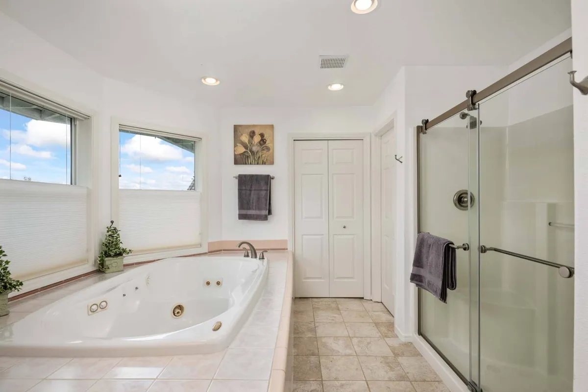 A serene bathroom with a jacuzzi tub, large windows with natural light, framed artwork, and a glass shower. The space invites relaxation and self-care.