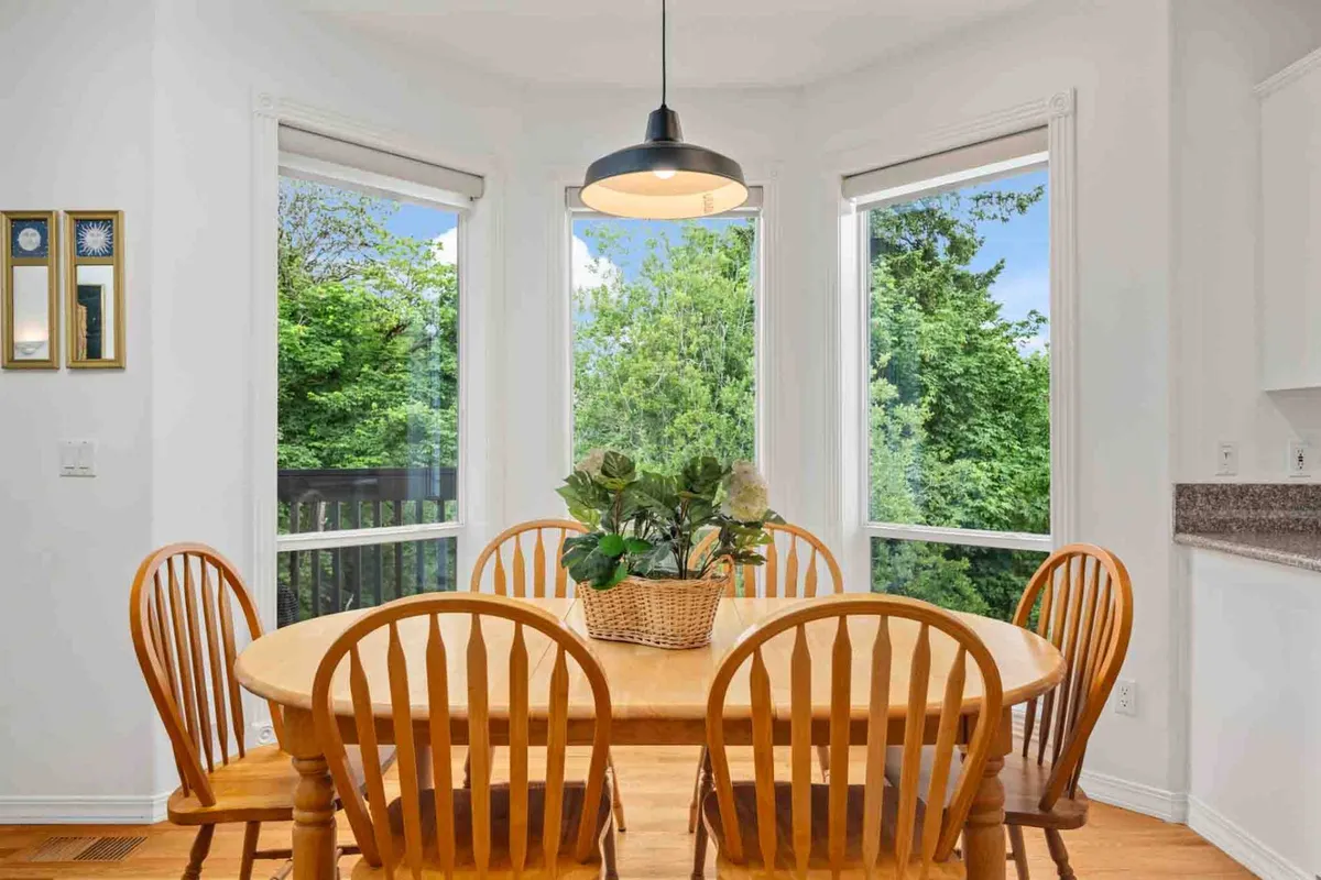 A warm, inviting dining area centered around a wooden table with surrounding chairs and a lush plant as a centerpiece. Large windows frame greenery outside, making the room feel connected to nature and perfect for group meals or quiet coffee mornings.