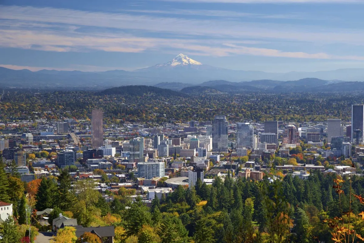A sweeping panoramic view featuring Mount Hood rising behind downtown Portland, framed by a lush canopy of trees and the urban cityscape. The scene captures both the natural beauty and the proximity to outdoor adventures.