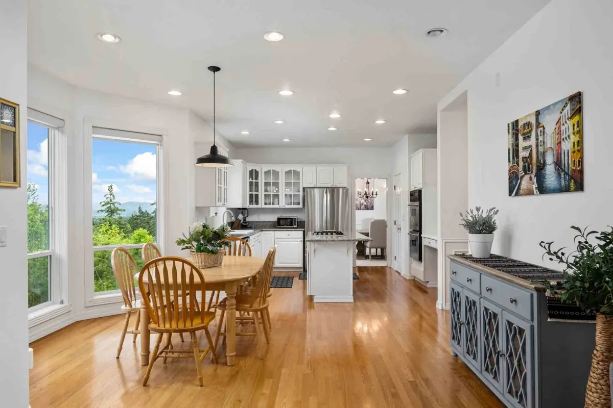 A cozy dining area adjacent to the kitchen, featuring a large dining table surrounded by chairs, expansive windows that let in natural light, and visible kitchen appliances in the background. The space is set for family meals and gatherings, with an inviting, homey atmosphere.