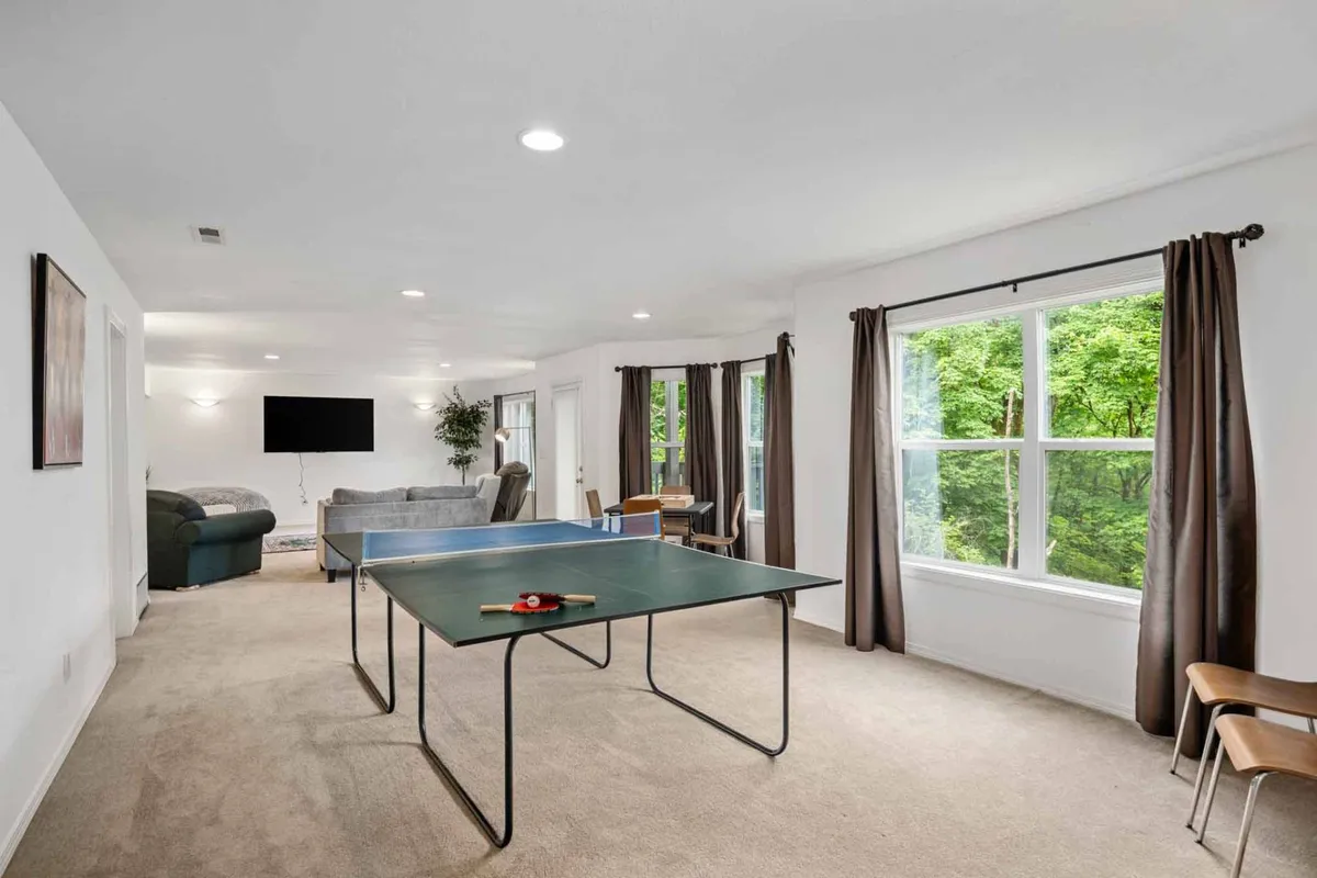 This image portrays a vibrant game room featuring a central ping pong table, large windows allowing natural light to flood in, a cozy seating area, and a television. The overall atmosphere is playful and inviting, capturing the essence of a dedicated entertainment space where guests can gather for various activities.