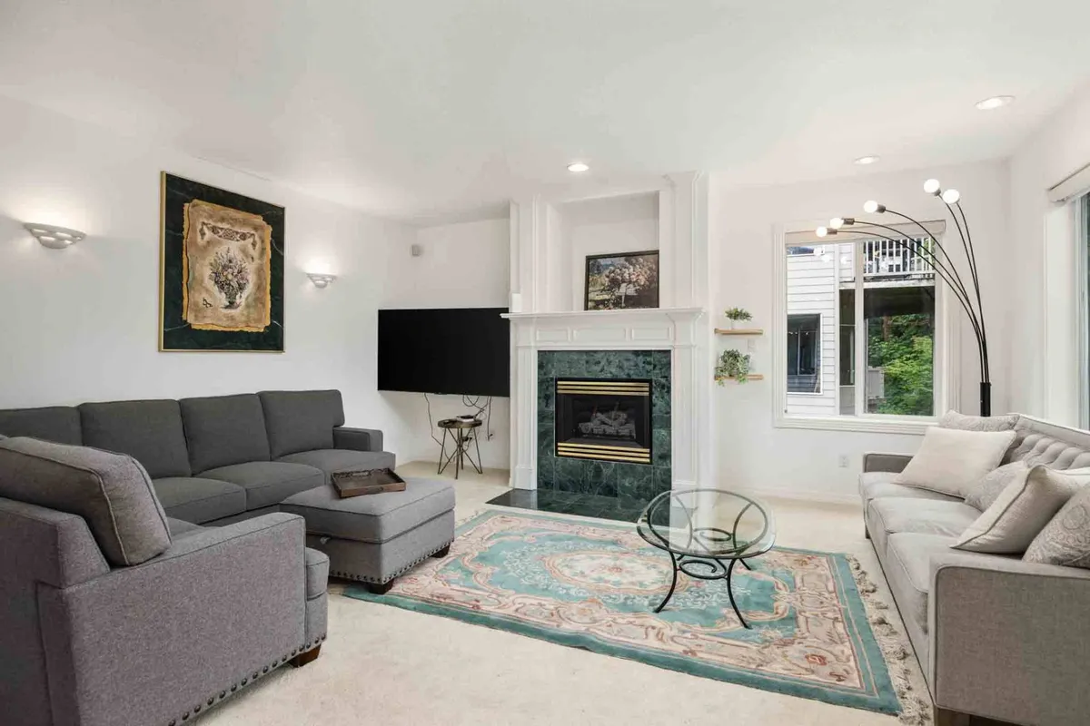 A welcoming living room with a stylish sofa set arranged around a modern fireplace and a glass coffee table, complemented by decorative artwork. Large windows flood the space with natural light and offer calming views of the surrounding greenery.