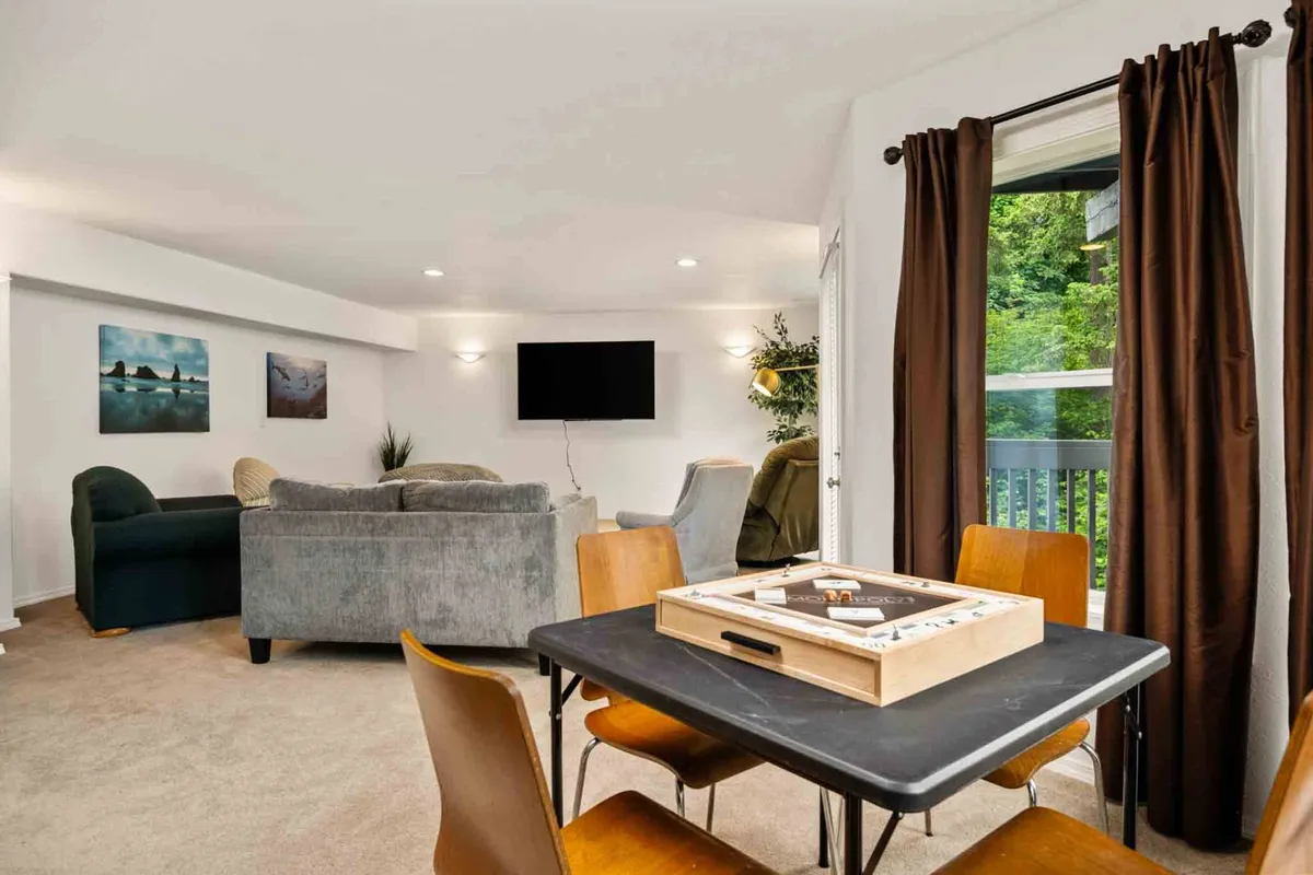 A comfortable seating area with a dining table in the foreground set up for a game, surrounded by a large window letting in natural light. The scene suggests relaxation, conversation, and shared moments, ideal for family game nights or connecting with friends.