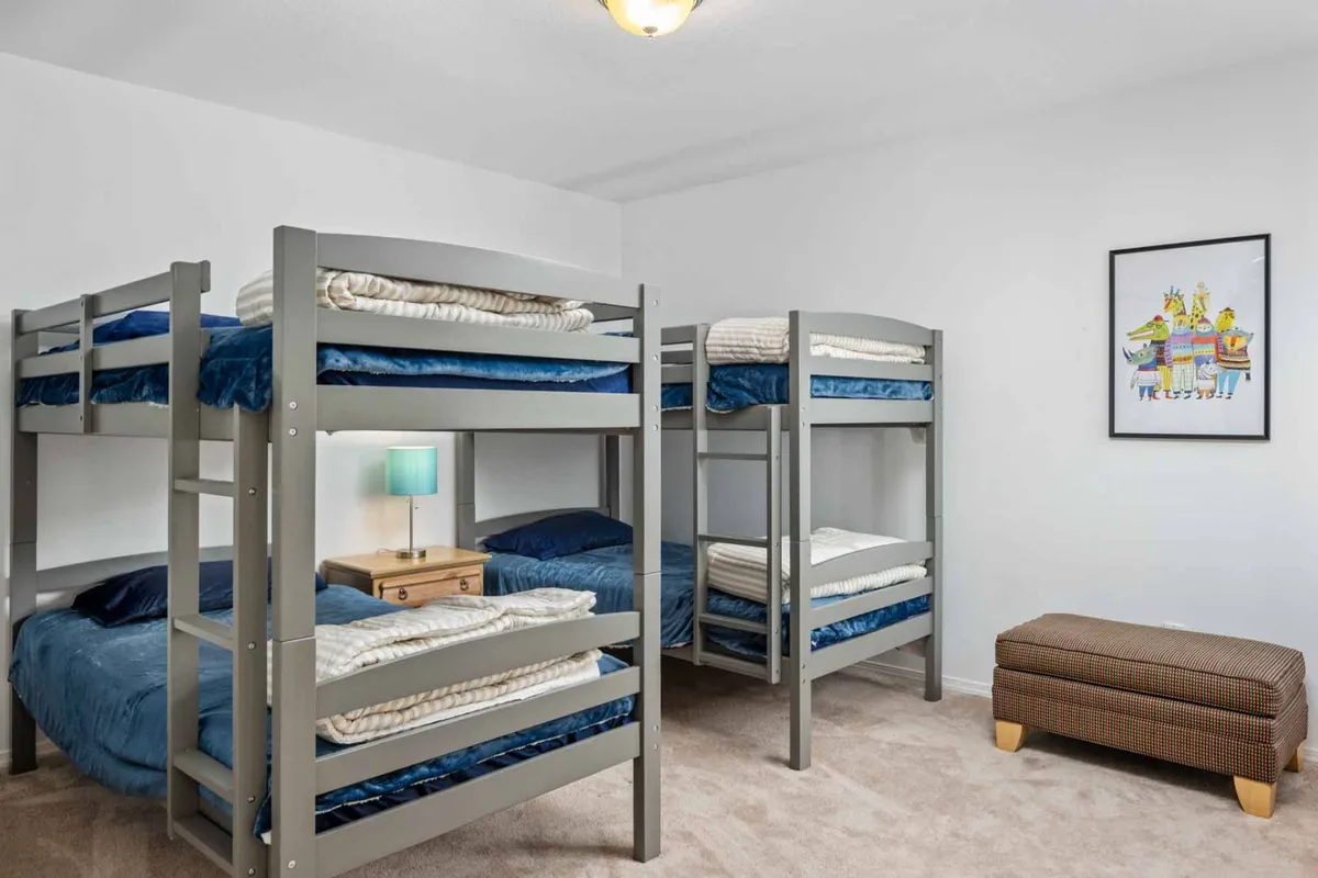A playful bedroom featuring twin bunk beds, colorful artwork, a nightstand with a lamp, and an ottoman. The layout is perfect for kids or groups, highlighting flexible sleeping arrangements.