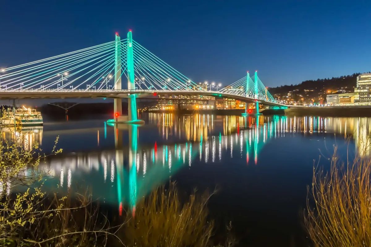 This exterior image features an illuminated bridge over water, with city lights and reflections in the background. The vibrant scene suggests nearby urban attractions and the lively atmosphere of the Tigard area in the evening.